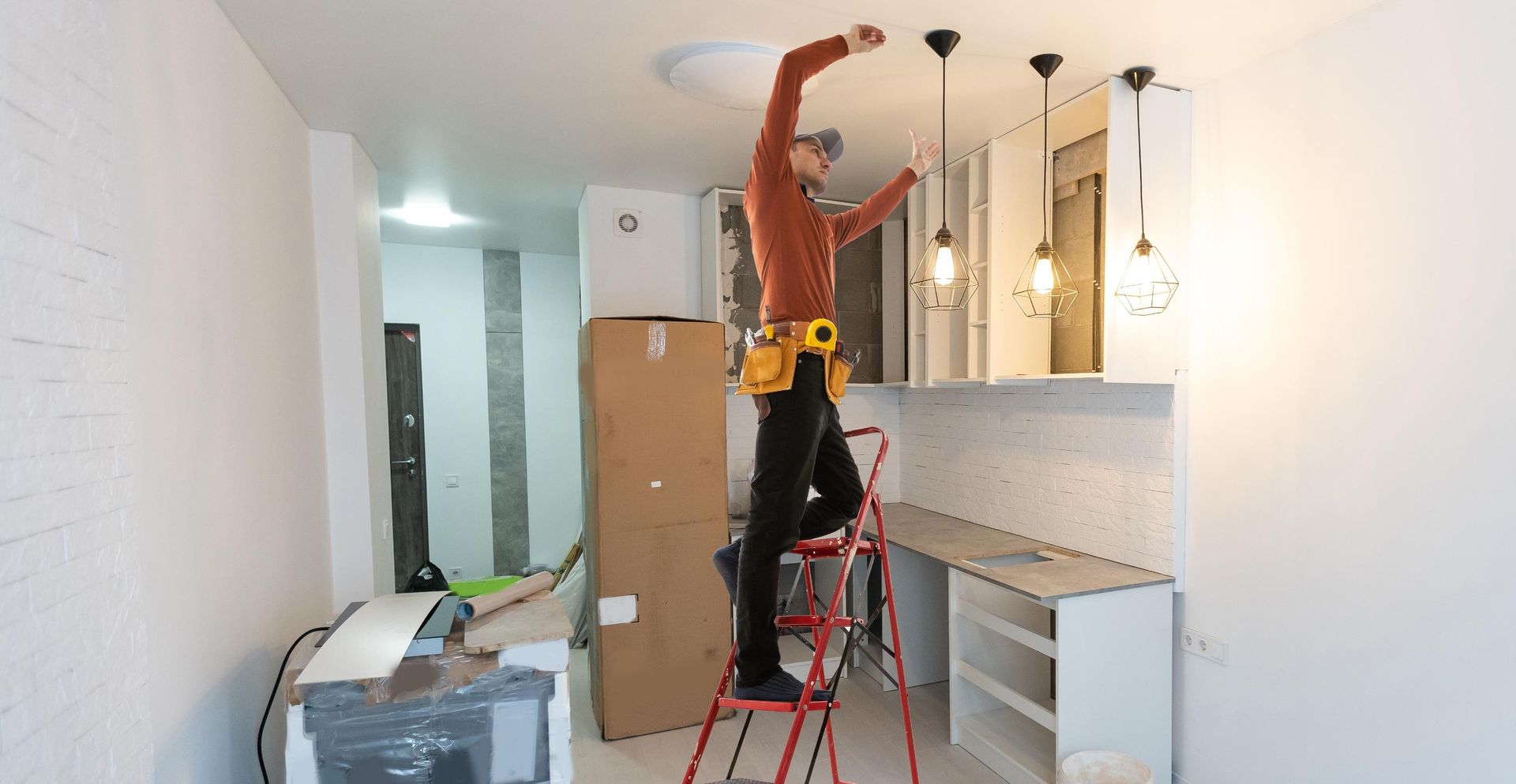A man is standing on a ladder fixing a light fixture in a kitchen.