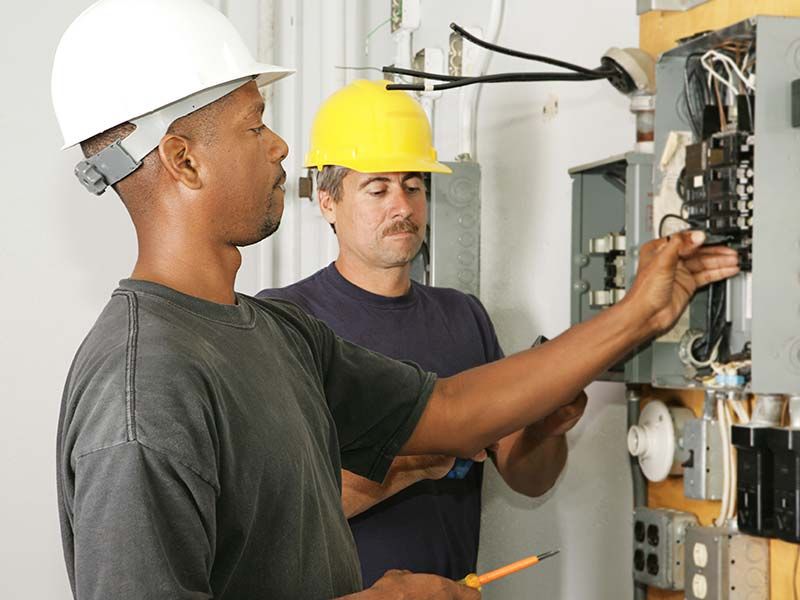 Two men wearing hard hats are working on an electrical box.