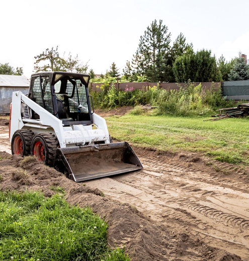White skid-steer loader digging a dirt path beside a grassy yard and white building.