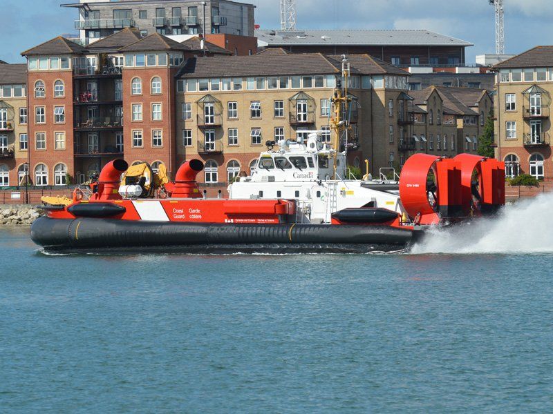 Orange rescue hovercraft on water near waterfront buildings, spraying water as it moves.