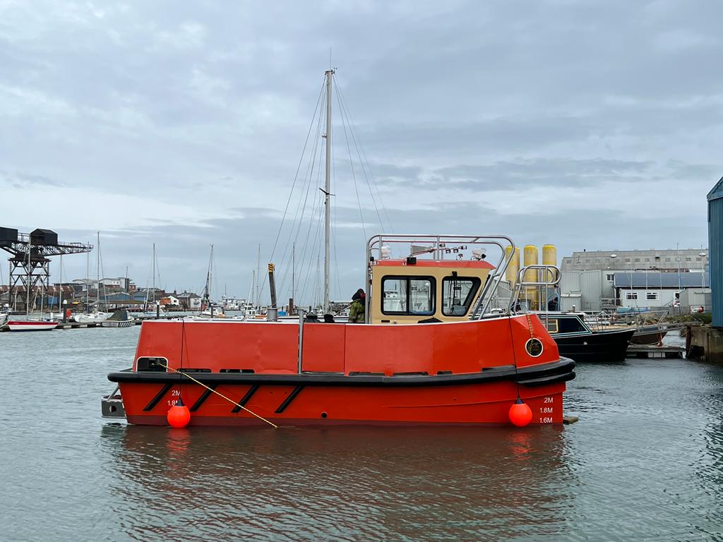Orange workboat floating in a harbor under a cloudy sky, with docked buildings in the background