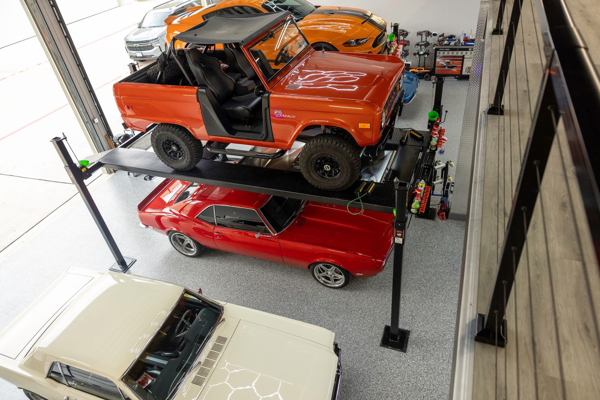 Cars in a garage: orange Bronco on a lift above a red muscle car, white car in foreground. | Auto Motive
