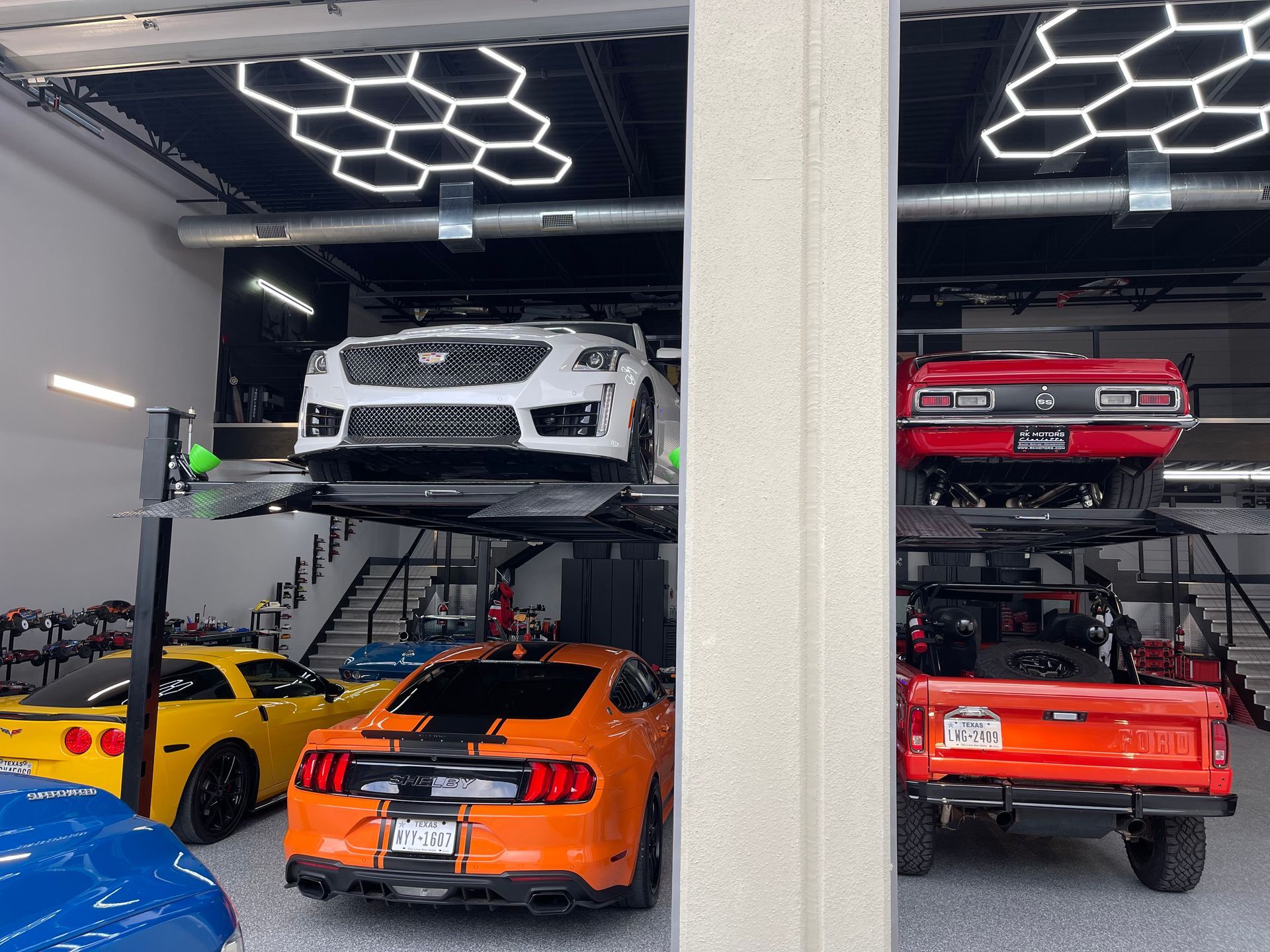 Garage with multiple cars, including a white Subaru and red Camaro on car lifts, and a yellow Corvette and orange Mustang below. | Auto Motive