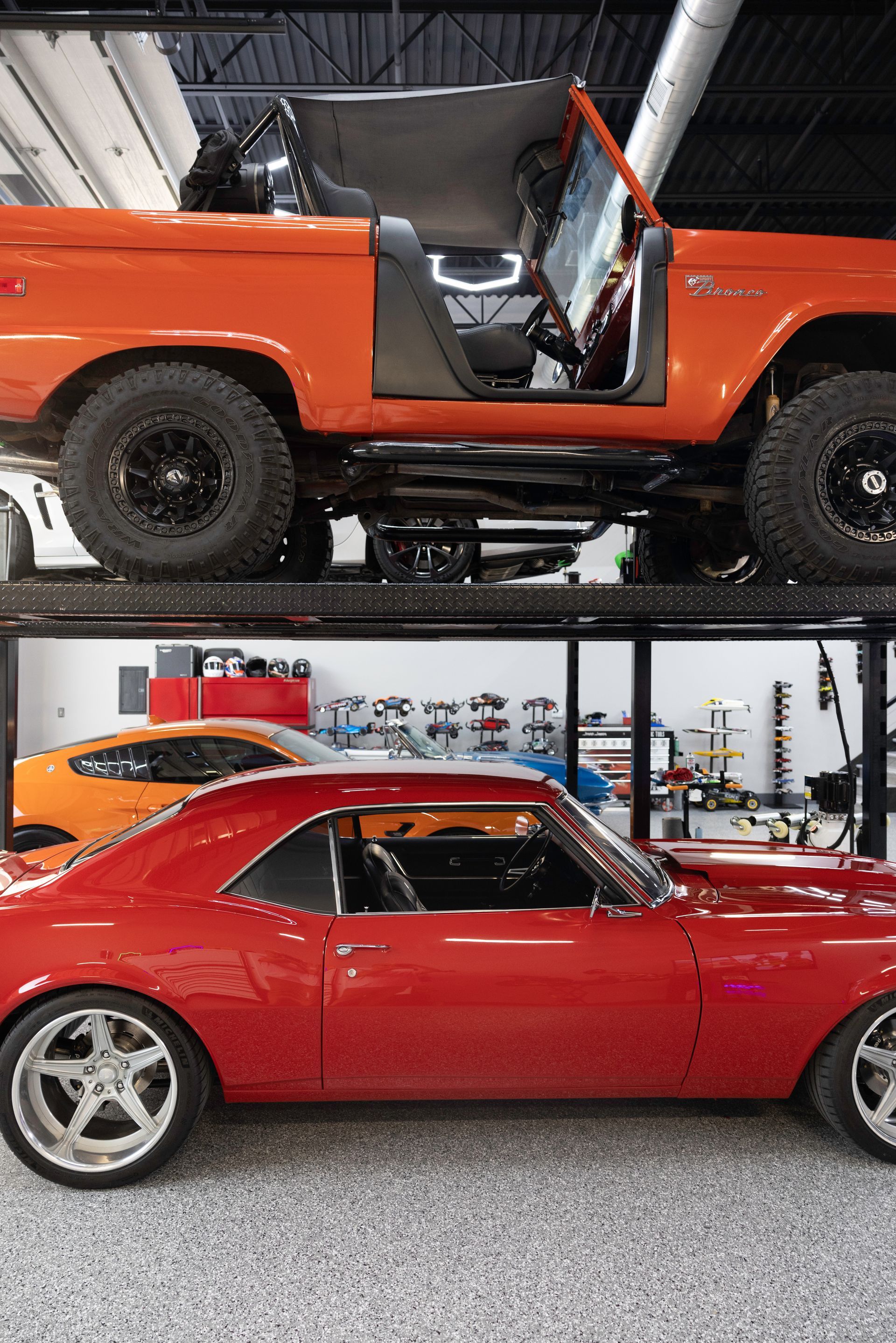 Two classic cars stacked. Orange Bronco on top, red coupe below. Garage setting. | Auto Motive