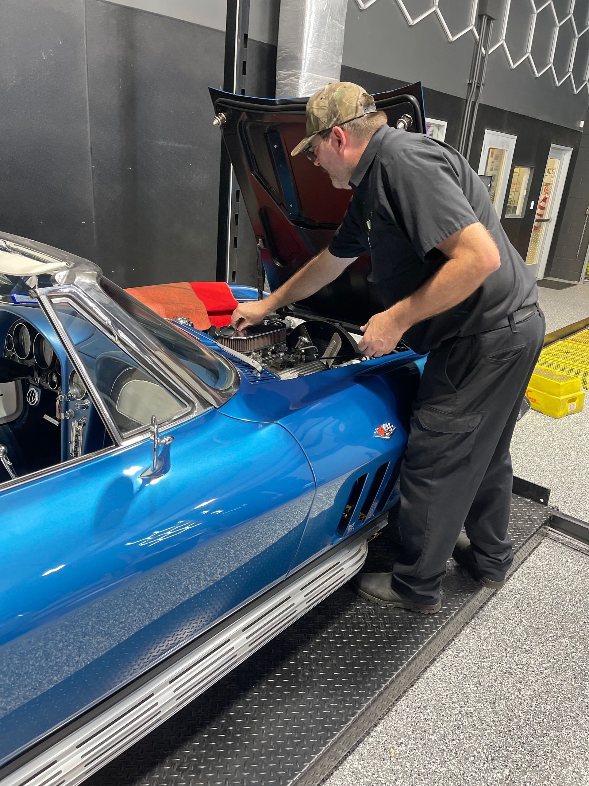 Mechanic working on a blue classic Corvette in a garage. He is leaning over the engine, inspecting it. | Auto Motive