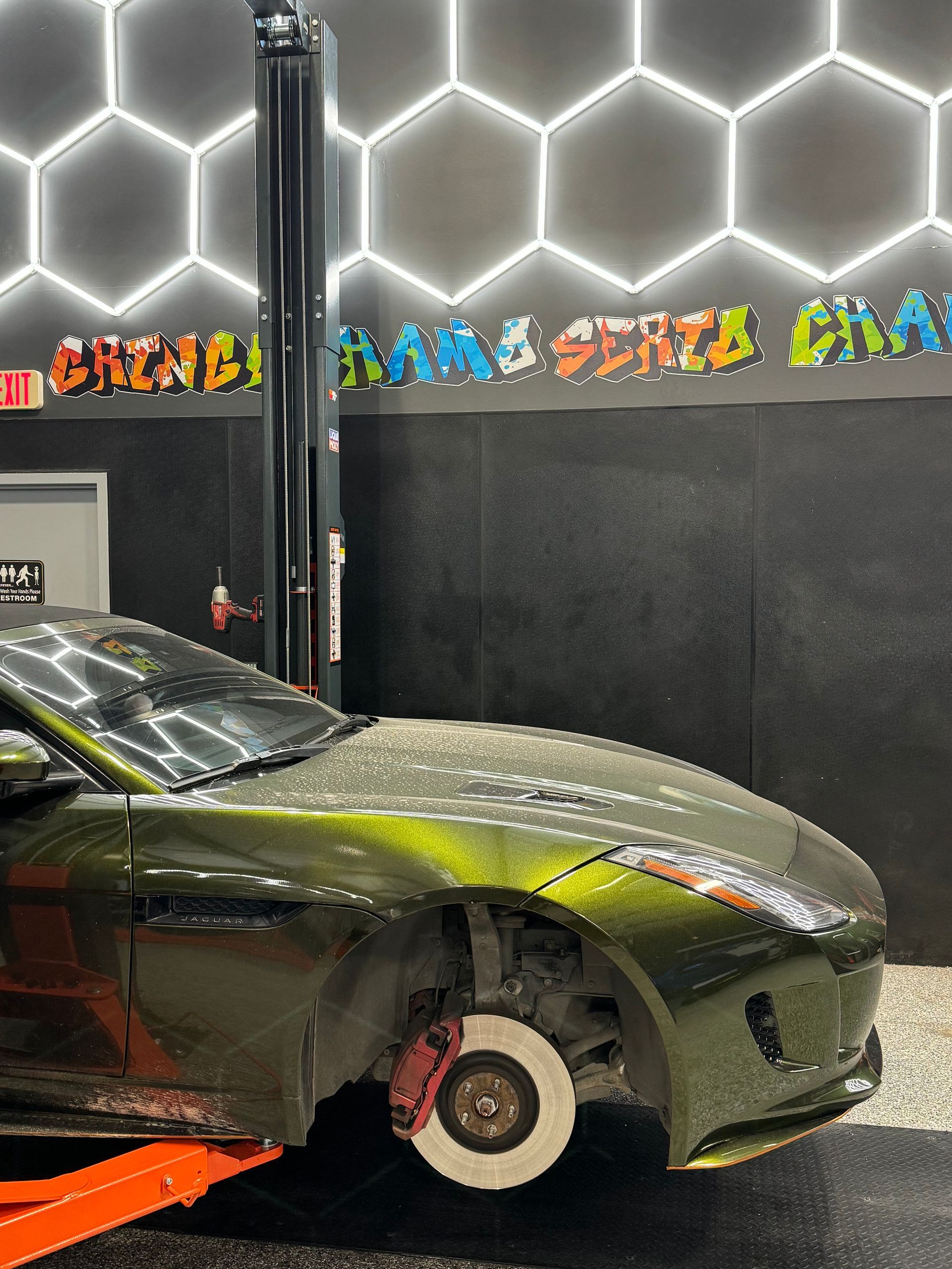 Green car on a lift in a garage with a hexagon pattern on the wall and graffiti. | Auto Motive