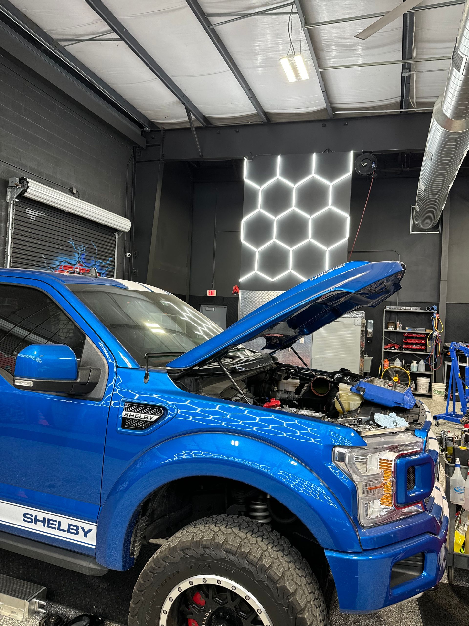 Blue Shelby Ford F-150 with open hood in auto shop, neon honeycomb lights in background. | Auto Motive