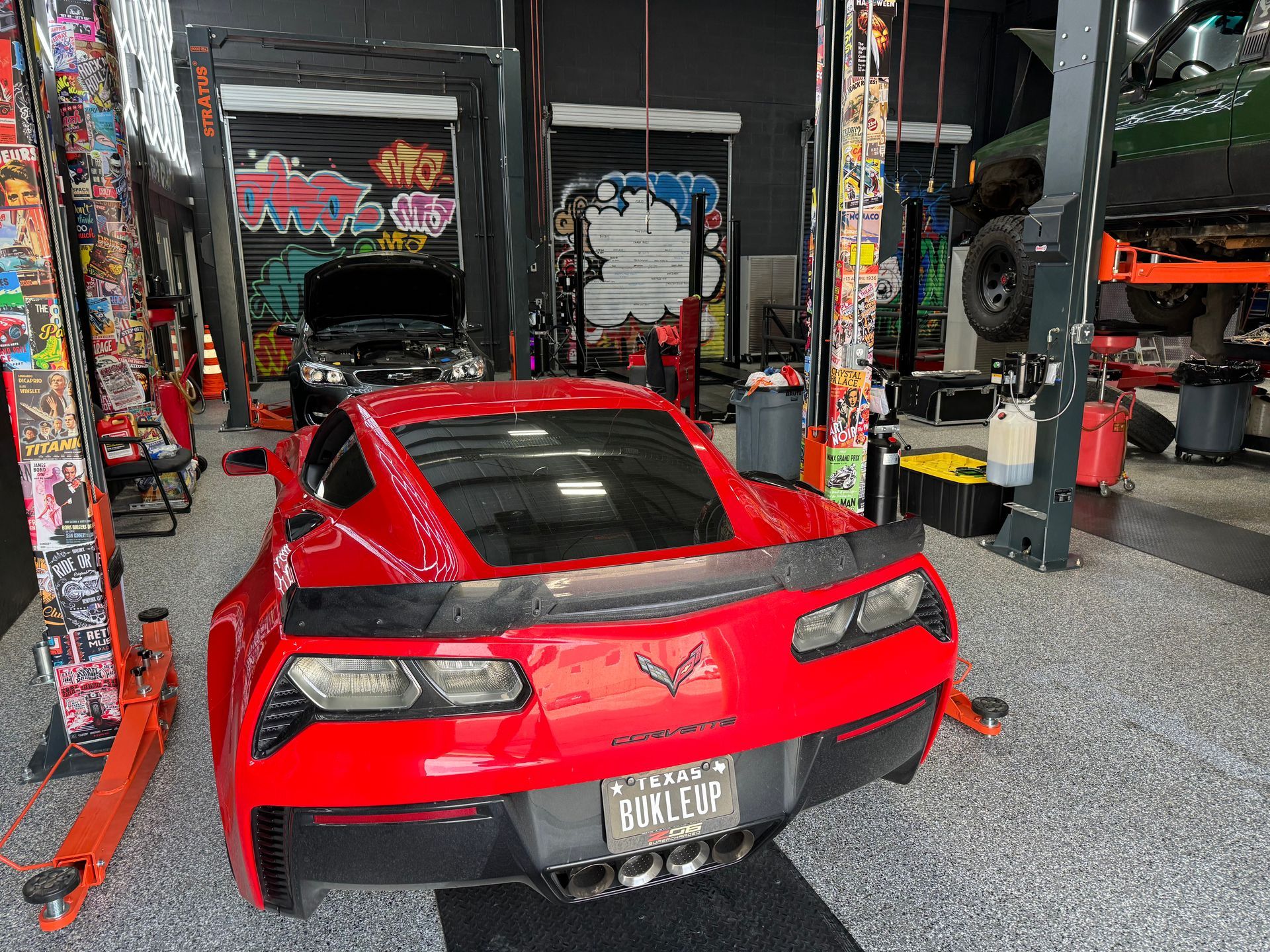Red Corvette in a garage, with lift and tools, surrounded by colorful wall art. | Auto Motive