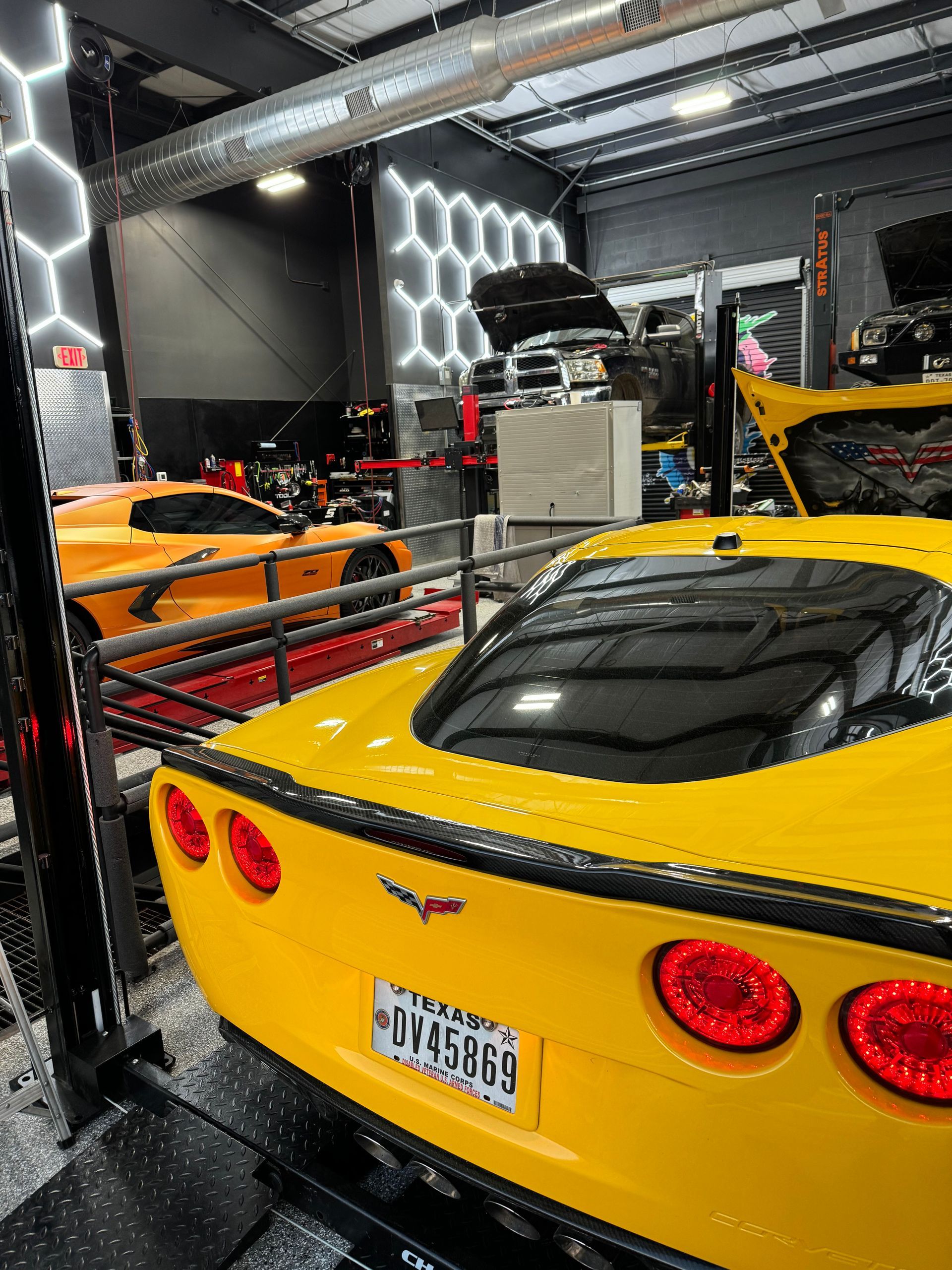 Yellow Corvette in a shop, another orange Corvette in background. Hexagon lights on the wall. | Auto Motive