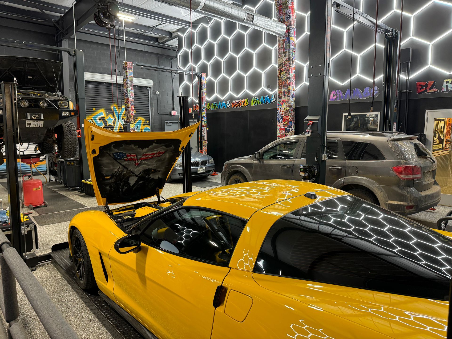 Yellow Corvette in a garage with hood open, other vehicles in the background. | Auto Motive