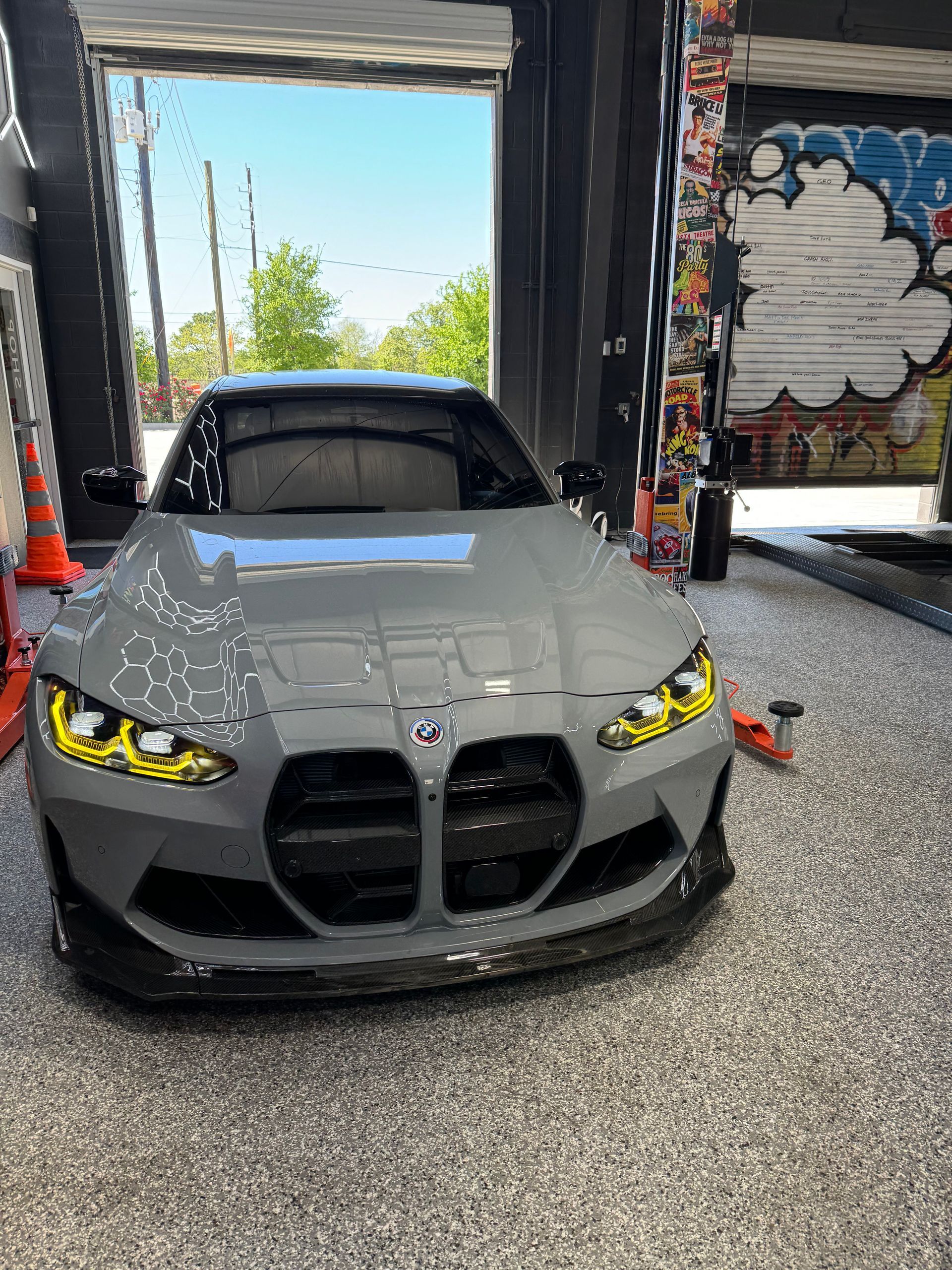 Gray BMW M4 with yellow headlights parked inside a garage; artwork on the right. | Auto Motive