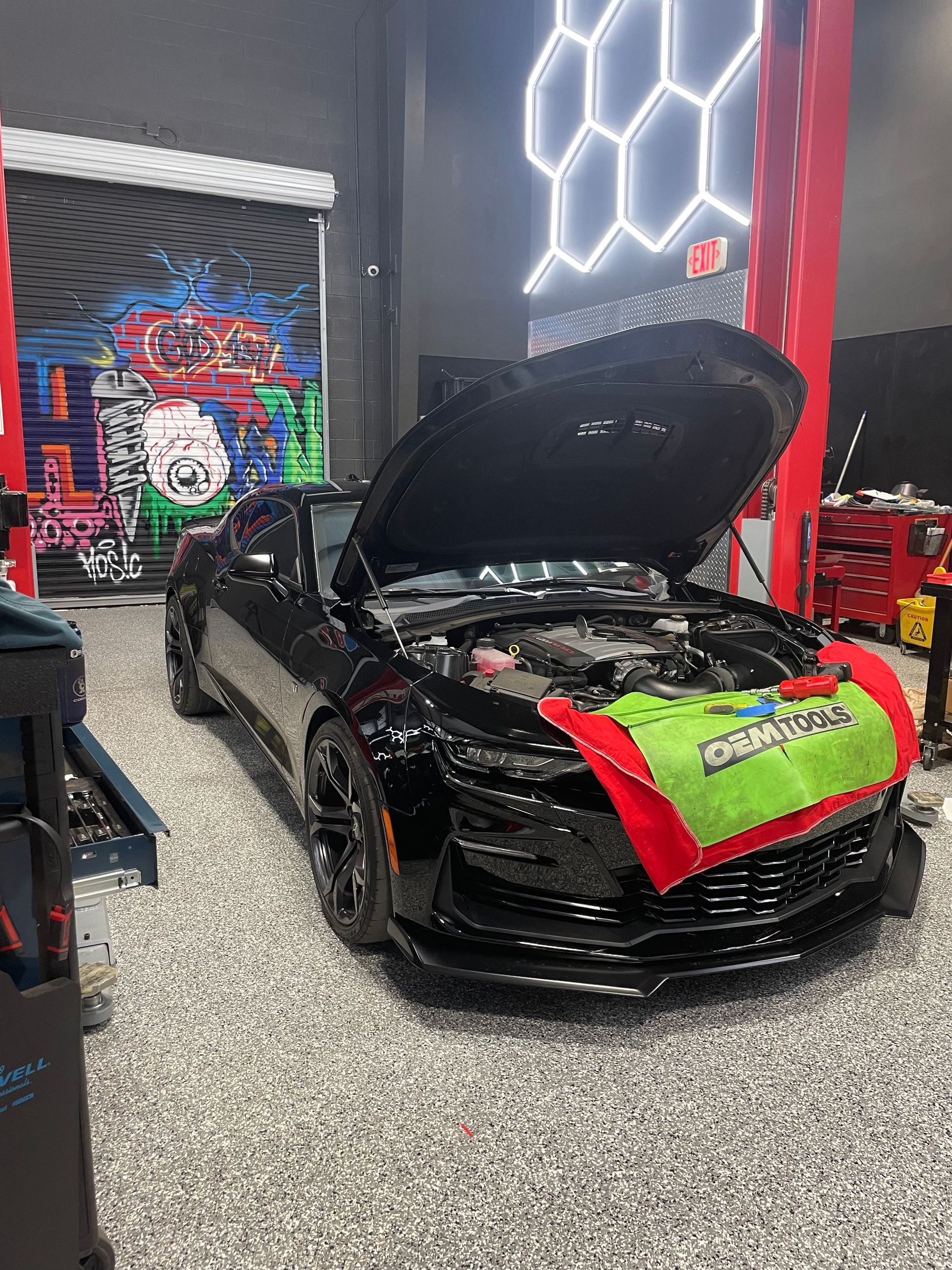 Black Camaro in a garage, hood open, with a red and green protective cover, and black rims. | Auto Motive