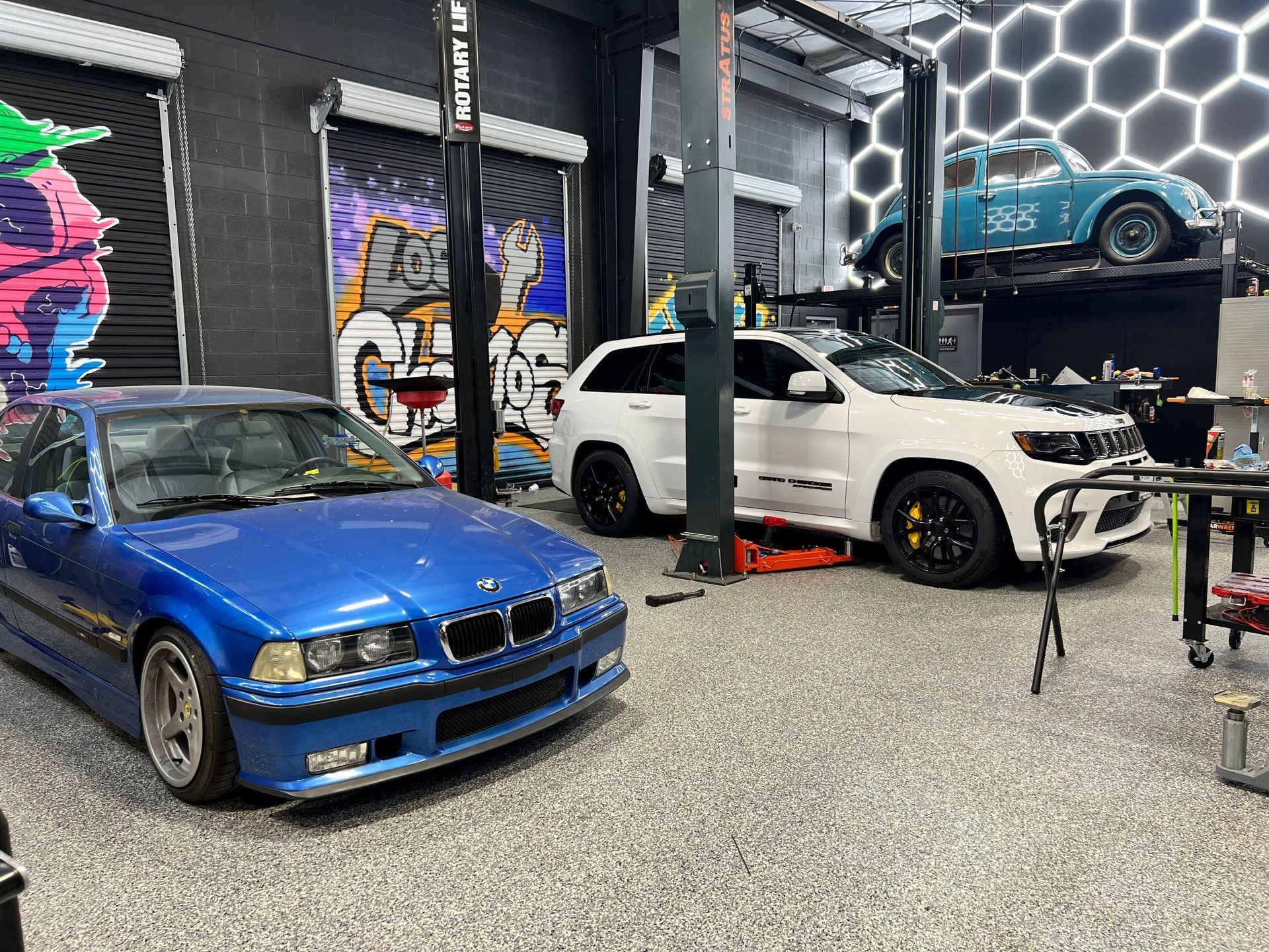 A blue BMW, white Jeep, and car on a lift inside a garage with graffiti and hexagon wall. | Auto Motive