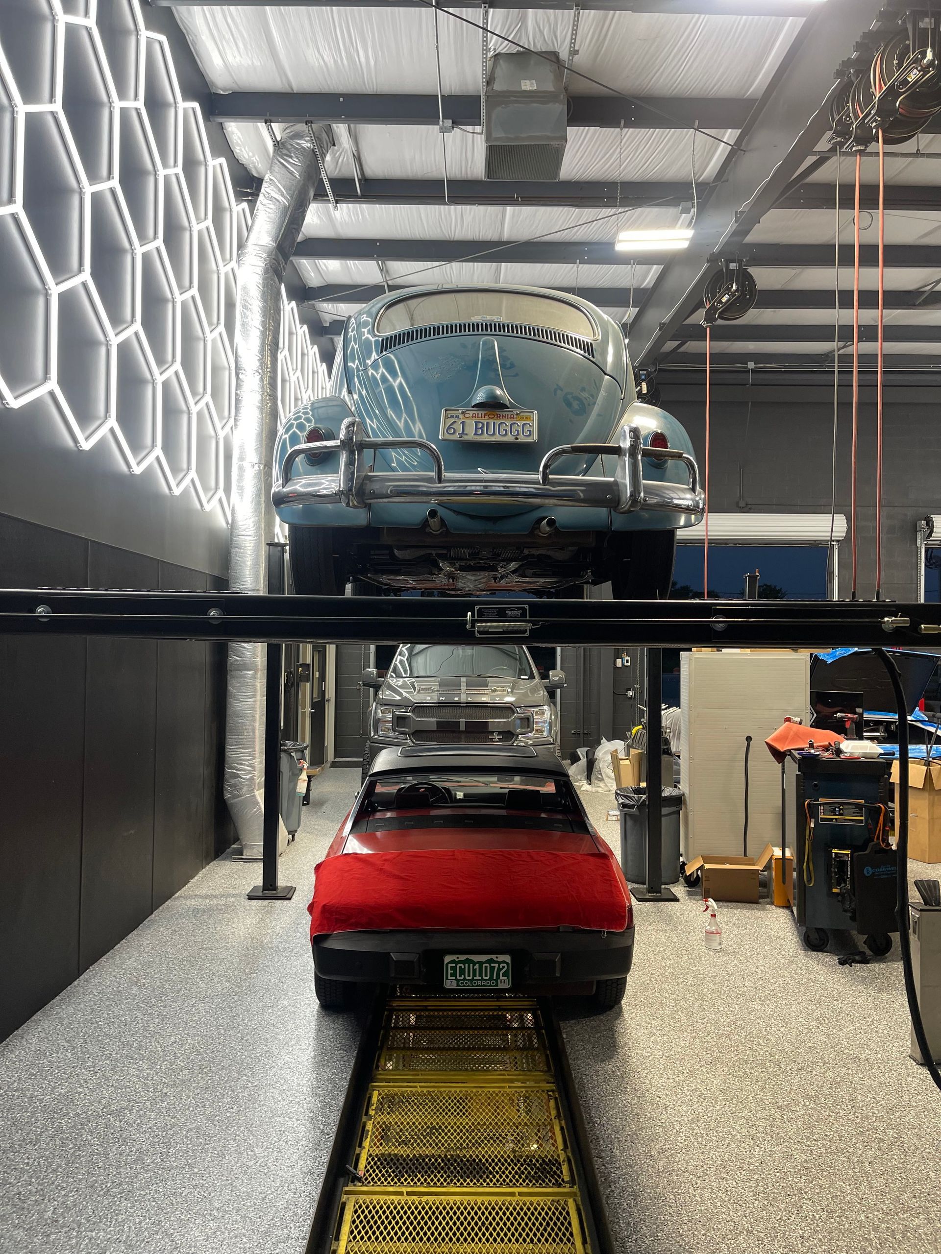 Two classic cars in a garage; red one on the ground, blue one elevated on a lift. Honeycomb wall. | Auto Motive