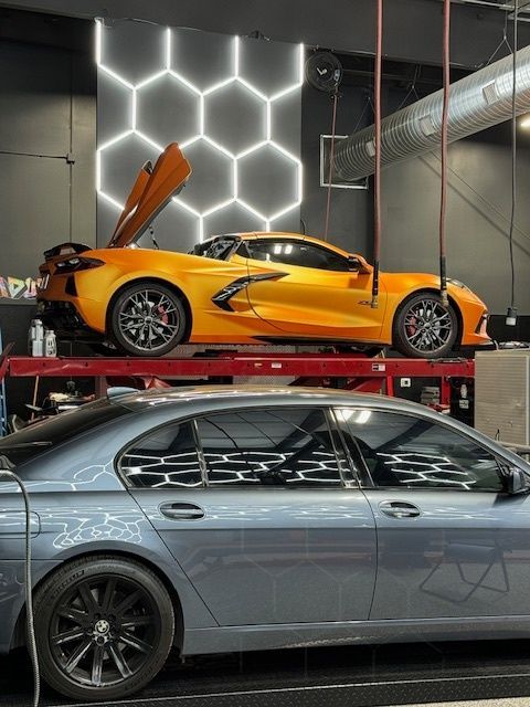 Orange Corvette elevated above gray sedan in a garage, with honeycomb-shaped wall lights.