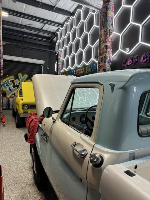Blue and cream pickup truck with hood up in a garage, yellow truck in background, hexagonal lit wall.