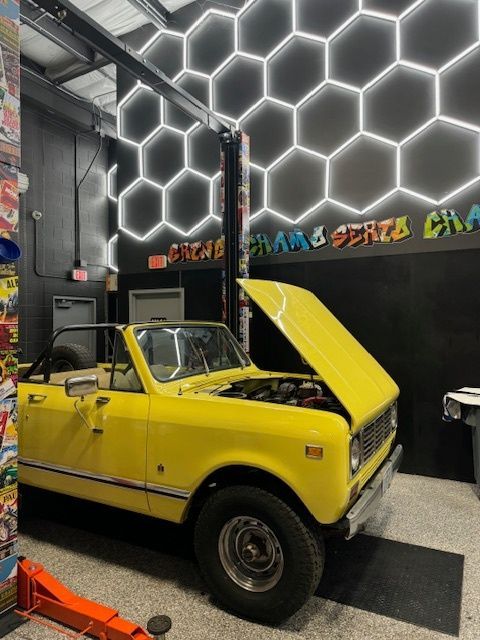 Yellow vintage convertible with open hood in a garage with honeycomb lighting.