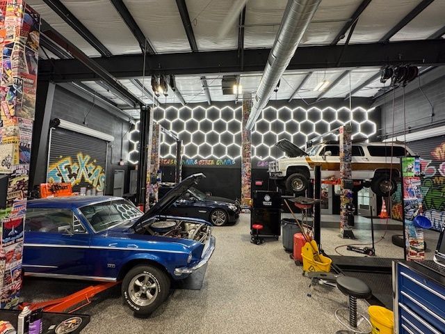 A well-lit auto repair shop with cars on lifts. A blue classic car has hood open, working on it.