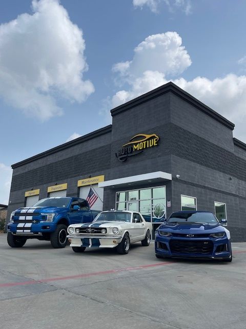 Three cars parked in front of a black building with a yellow sign. Blue truck, white Mustang, and blue Camaro. | Auto Motive