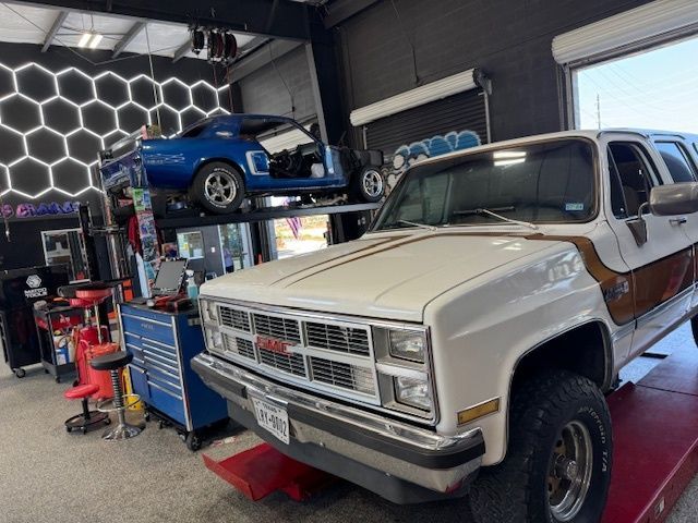 White and brown GMC Suburban in a garage, blue car on a lift, tools, and a honeycomb wall.