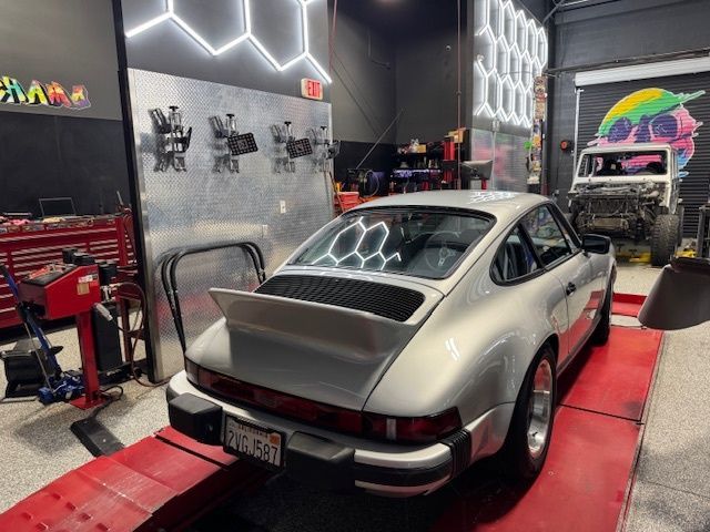 Silver Porsche sports car on a lift in a garage, with tools and a vehicle being worked on in the background.
