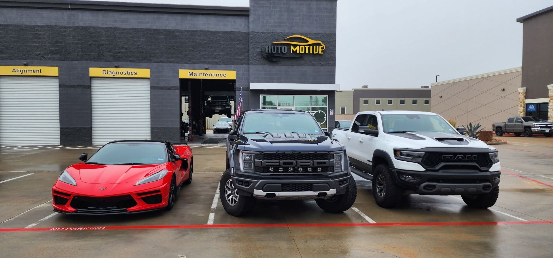 A red Corvette, black Ford Raptor, and white Ram TRX parked in front of a business on a wet day.