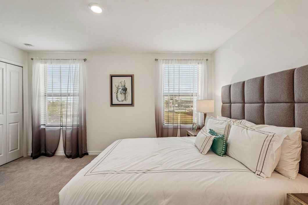 Bright bedroom with white bedding, beige headboard, dresser, and two windows with blinds