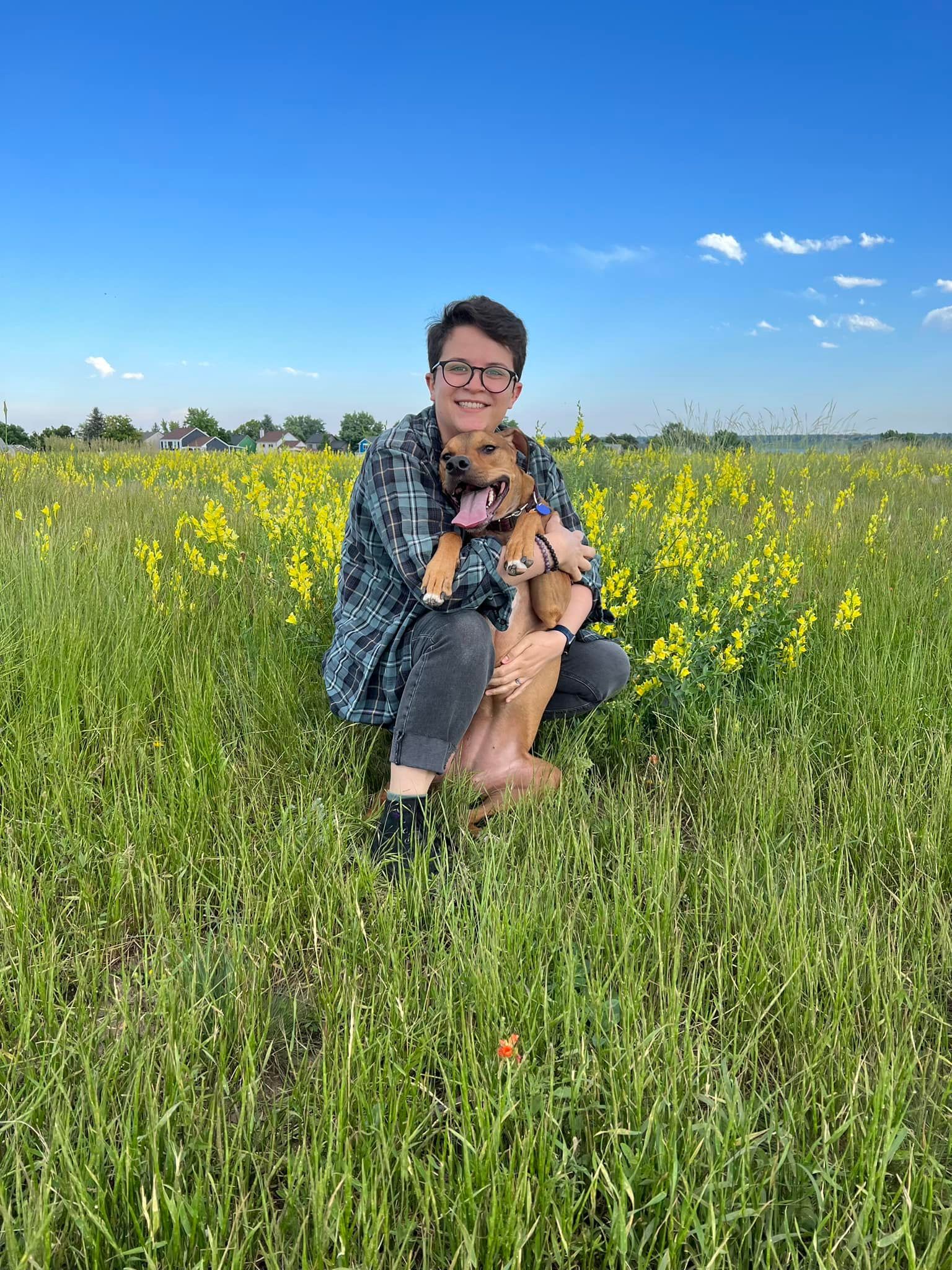 Person with glasses crouches in a field of yellow flowers, holding a small brown dog with its tongue out. Blue sky.