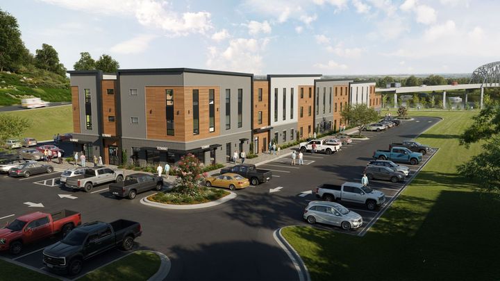 Multi-story commercial building with parking lot. Gray, brown, and white facade. Cars parked in front. Highway overpass visible in the distance.
