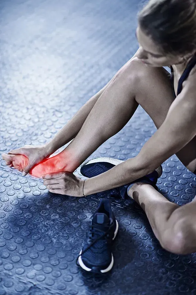 Woman holding her red-lit ankle while seated on a gym floor, with a running shoe nearby