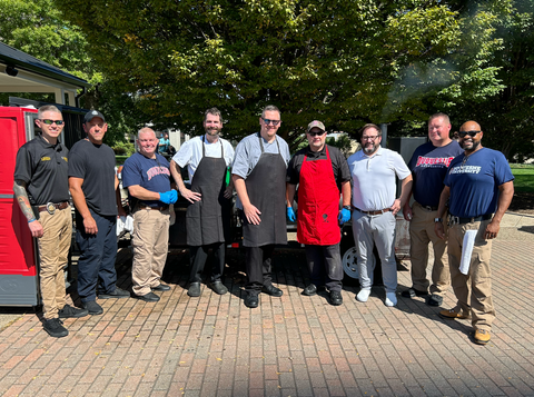 Ten people stand in a line outdoors in front of a tree, some wearing aprons, on a brick patio.