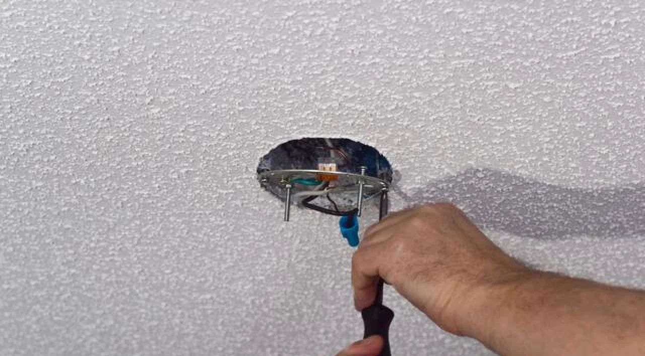 Hand repairing a ceiling light fixture with exposed wires in a white ceiling.