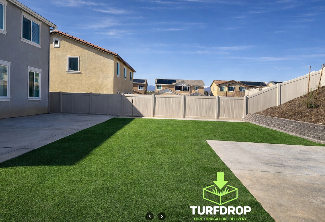 Backyard with green turf lawn, concrete patio, wooden fence, and neighboring houses under a blue sky