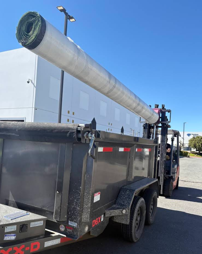 Forklift loading a long silver pipe onto a black trailer in an outdoor lot