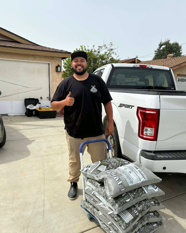 Man standing by a white pickup truck, giving a thumbs up beside stacked bags on a hand truck in a driveway