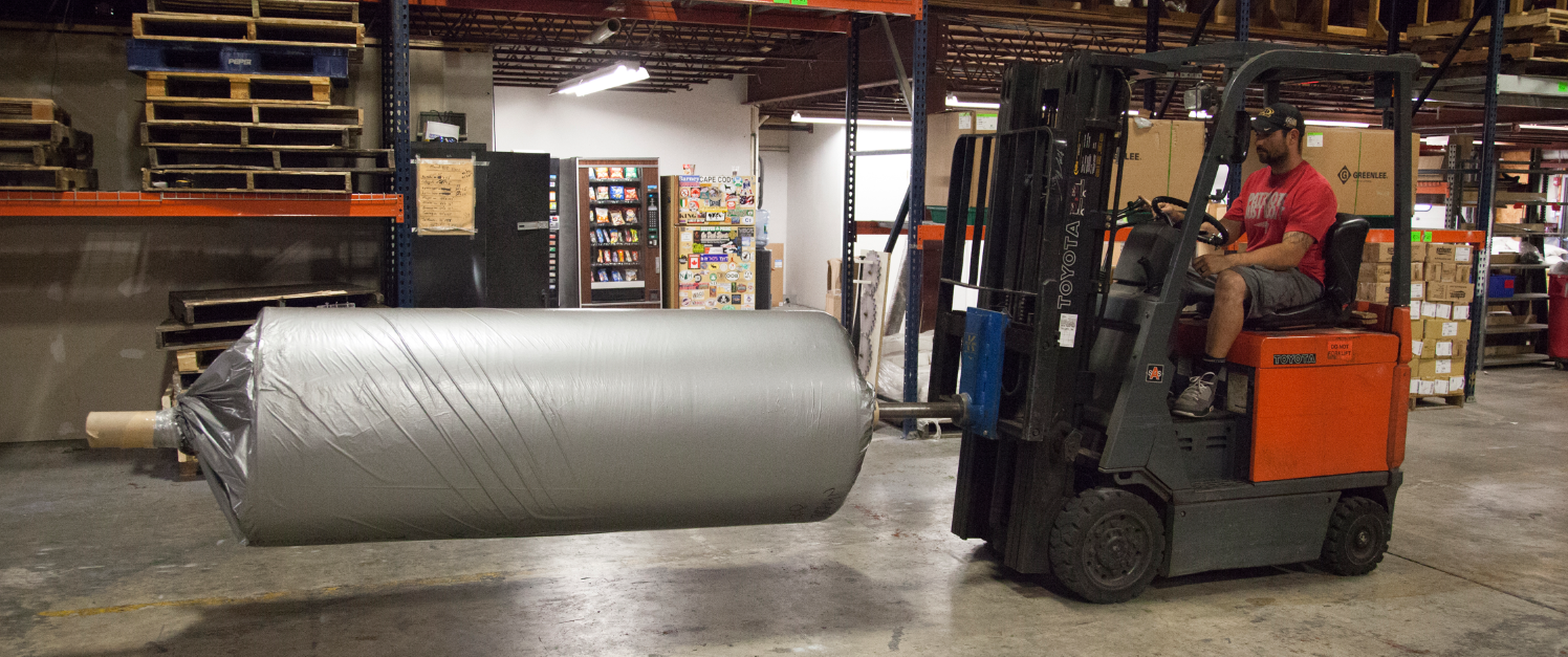 Forklift in a warehouse moving a large rolled silver sheet on the floor
