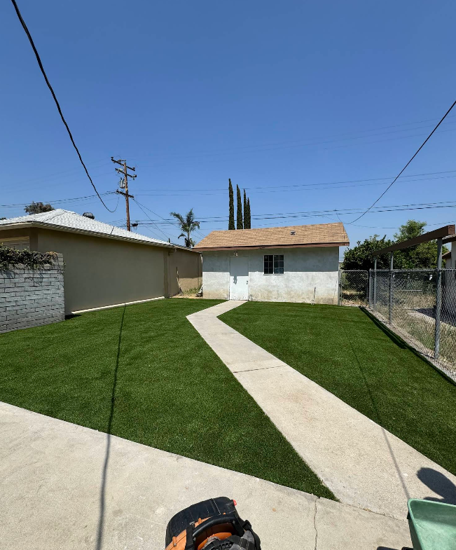 Backyard with green lawn, concrete paths, and small houses under a clear blue sky
