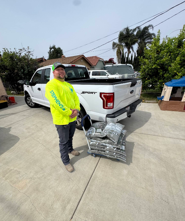 Person in neon yellow shirt stands by a white pickup truck with stacked packaged items on driveway.