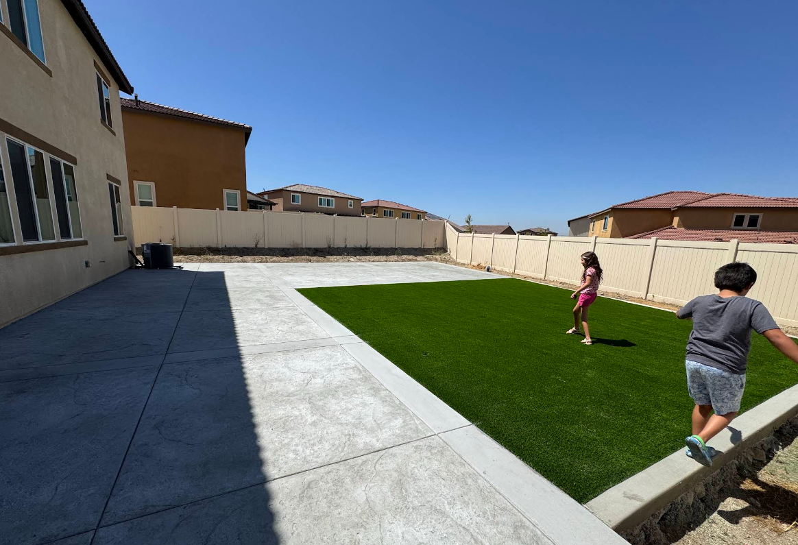 Backyard with artificial turf and concrete patio; two children running near a house under a blue sky