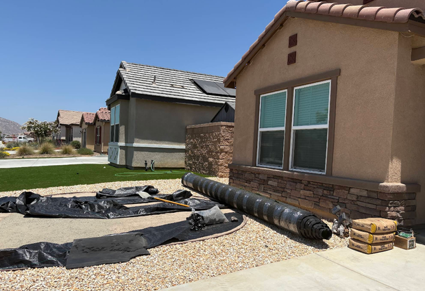 Homes under construction with roofing materials, gravel, and a tan stucco house in the foreground.
