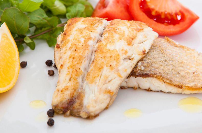 Filetto di pesce grigliato con limone, fette di pomodoro, prezzemolo e grani di pepe su un piatto bianco.