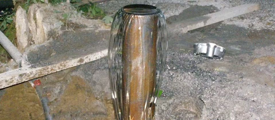 Un tuyau métallique rouillé, dressé verticalement sur un sol rocailleux, près d'un petit objet blanc.