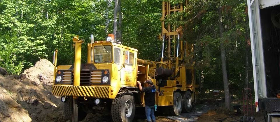 Un camion utilitaire jaune et des ouvriers sur une route forestière bordée de grands arbres, avec du matériel lourd à l'arrière.