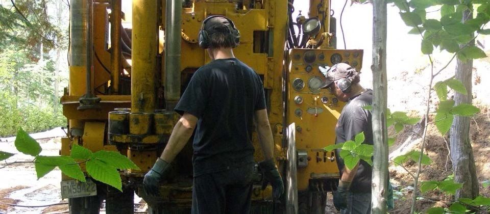Des ouvriers manipulent des engins de forage jaunes dans une zone boisée