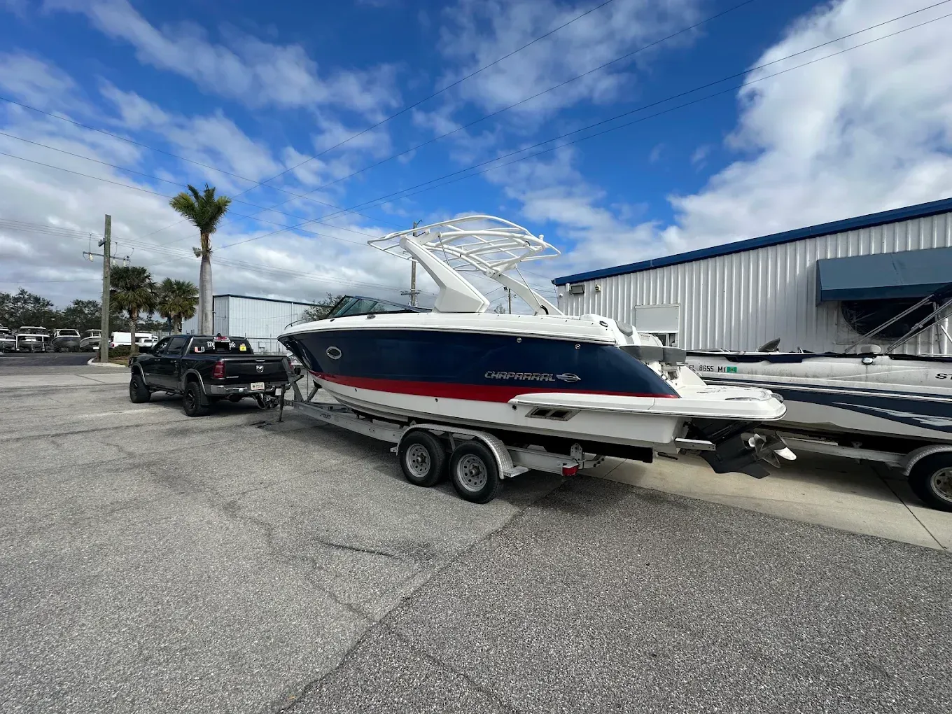 Boat on a trailer, blue and white hull with red stripe, parked in a lot on a sunny day with a truck nearby.
