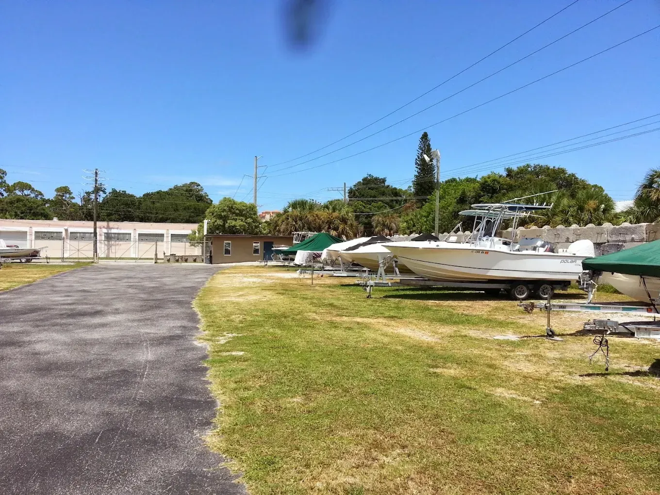 Boats on trailers in a grassy lot next to a paved area. Blue sky, sunny day.