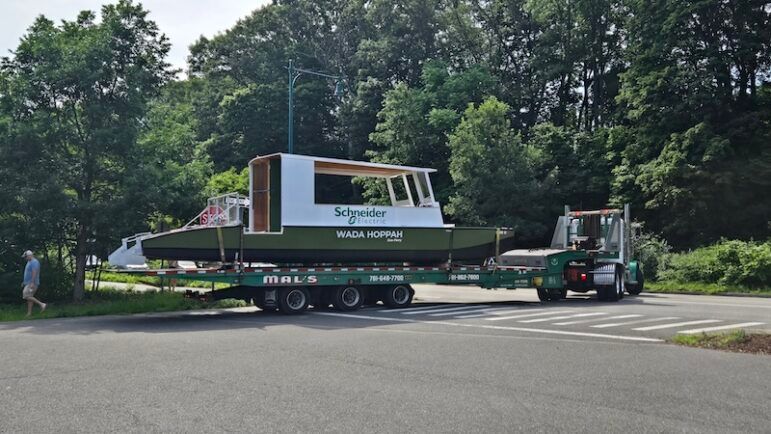 Wada Hoppah's electric ferry Just Get On loaded on a flatbed trailer, being transported by truck to the Charles River for launch.