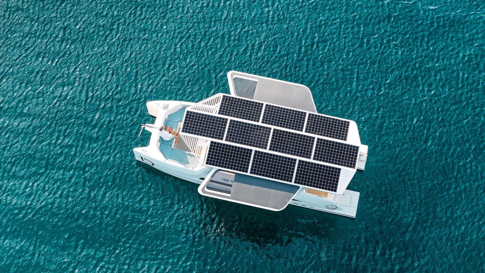 Birdseye view of solar panels on catamaran boat in the middle of green/blue water. There is one person seen sunbathing at the front of the boat