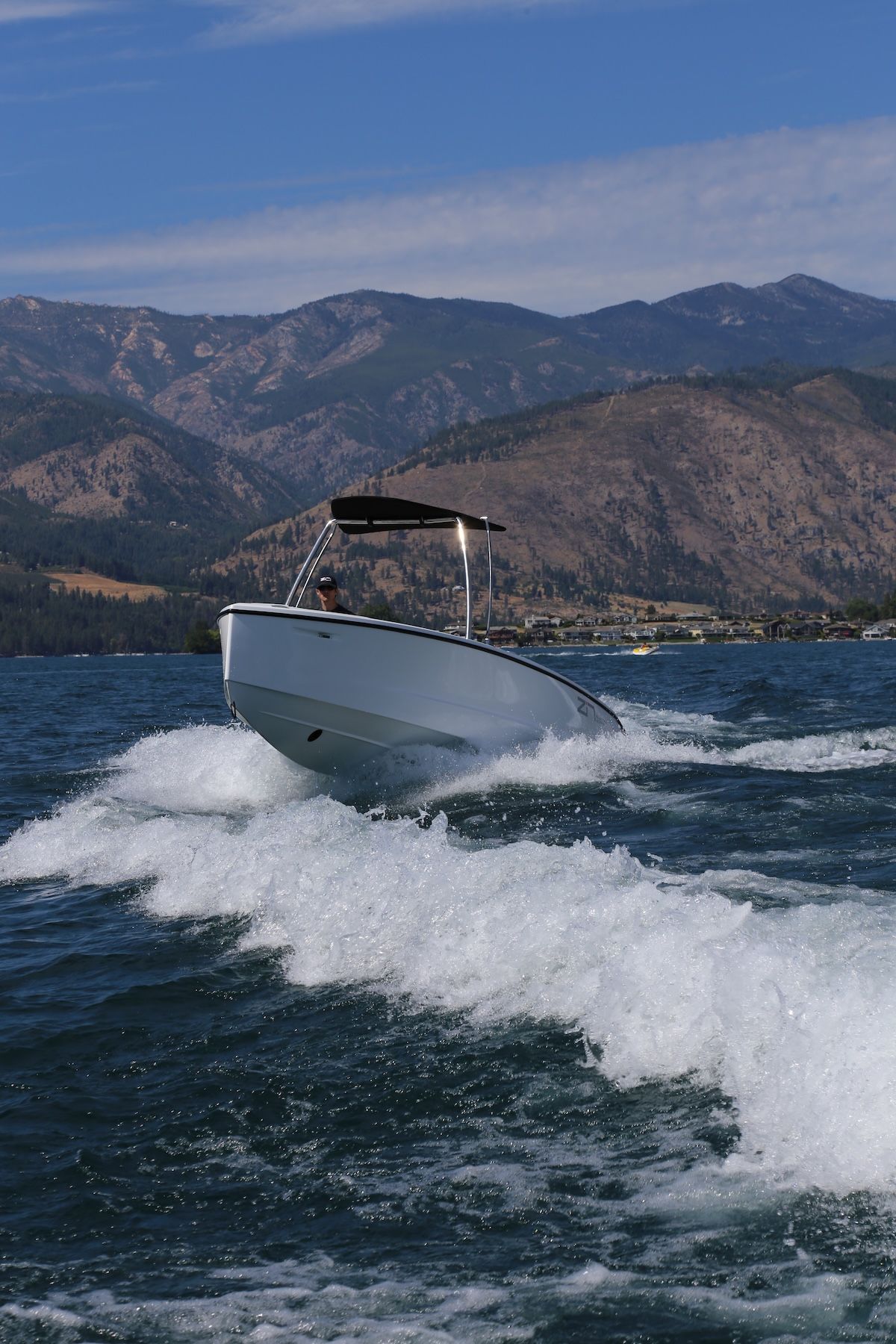 white boat going at high speeds through wake in a lake with mountains in the background. It has a T Top that is black