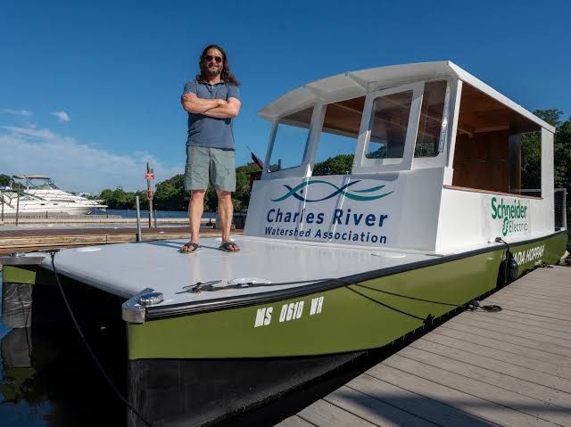 Drew Rollert stands with arms crossed on the deck of Just Get On, Wada Hoppah's green-and-white electric ferry, docked on the Charles River